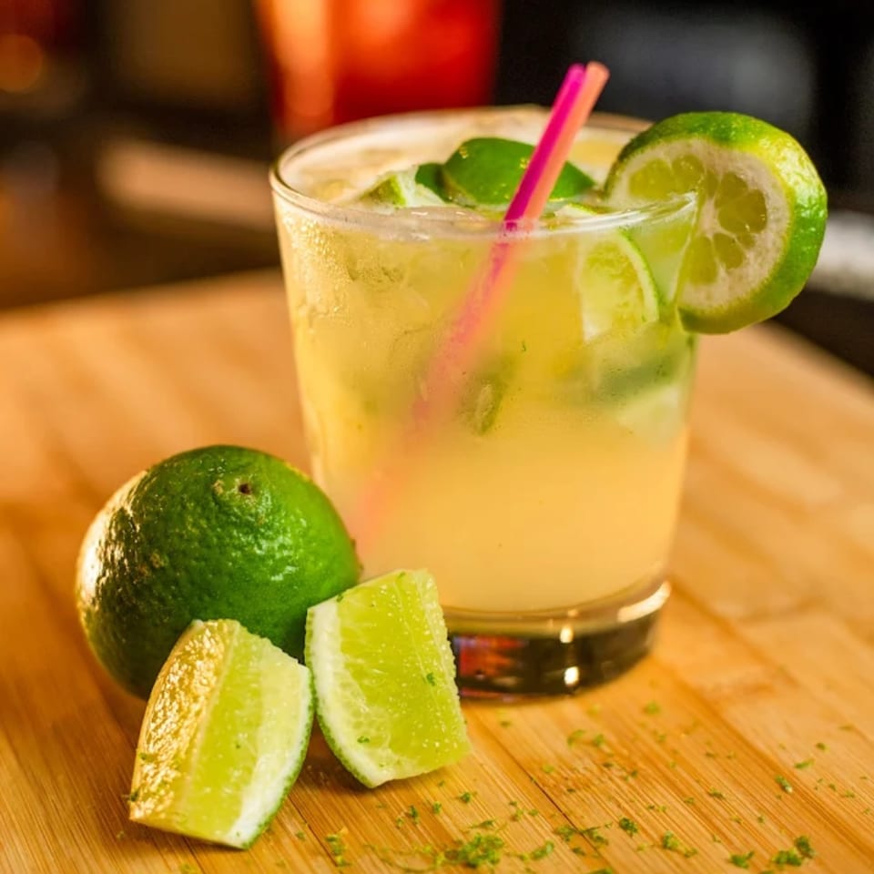 A refreshing caipirinha cocktail with lime slices and a pink straw on a wooden surface.