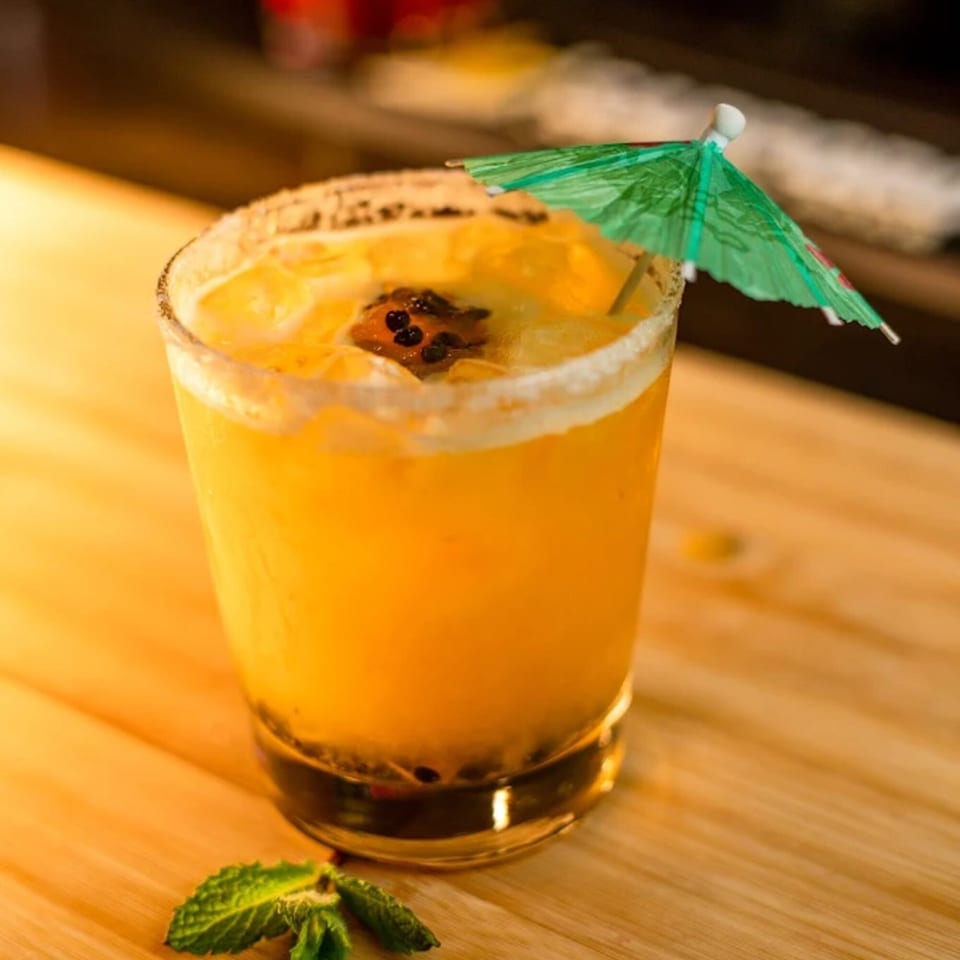 A vibrant orange cocktail with passion fruit seeds, a sugar-rimmed glass, and a green umbrella.