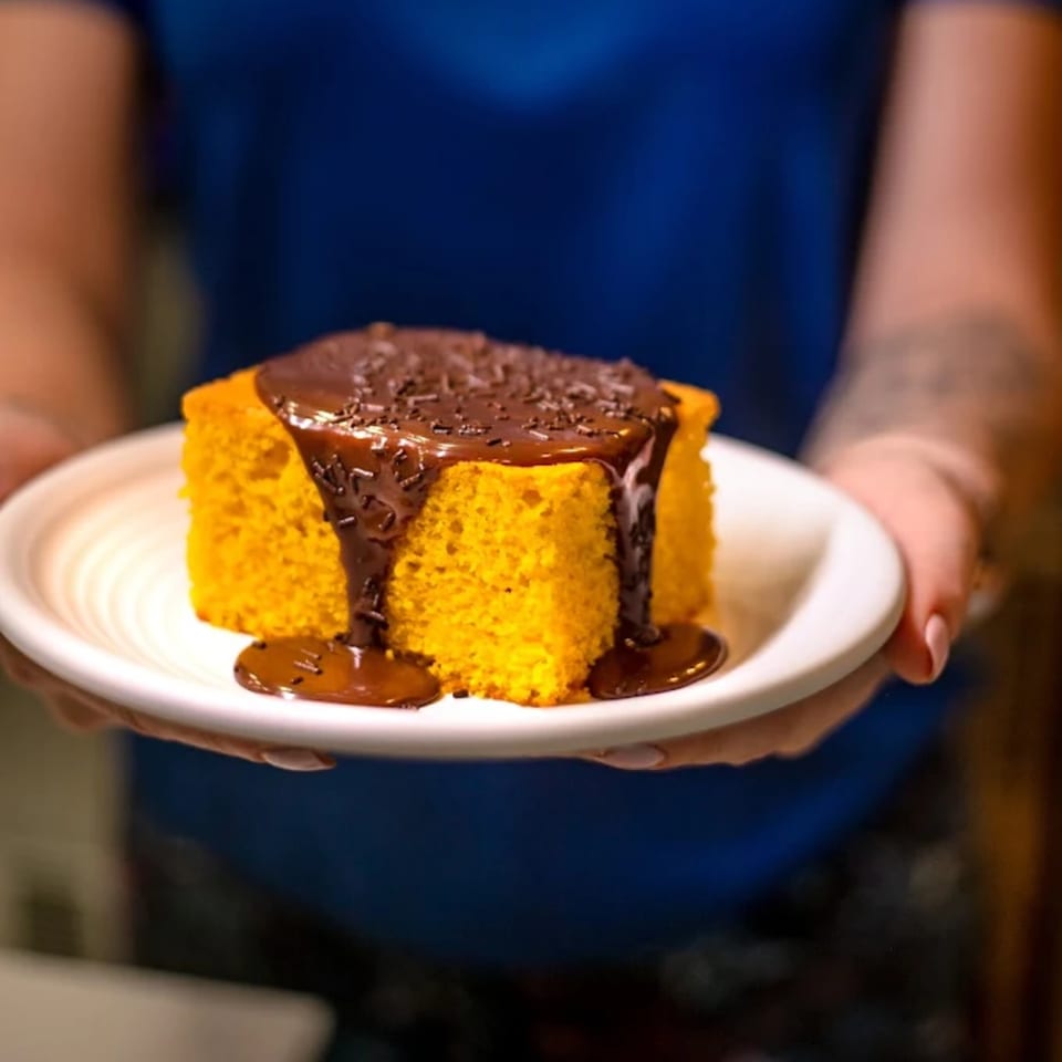 Hands holding a white plate with a slice of carrot cake topped with chocolate syrup and sprinkles.