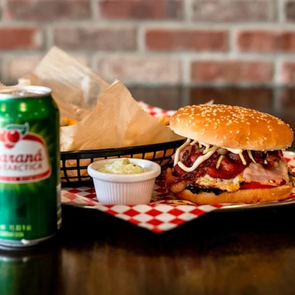A burger with bacon, egg, and sausage, fries, dip, and a green soda can on a checkered tray.