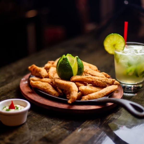 Fried fish pieces with lime, a dipping sauce, and a mojito on a dark table.