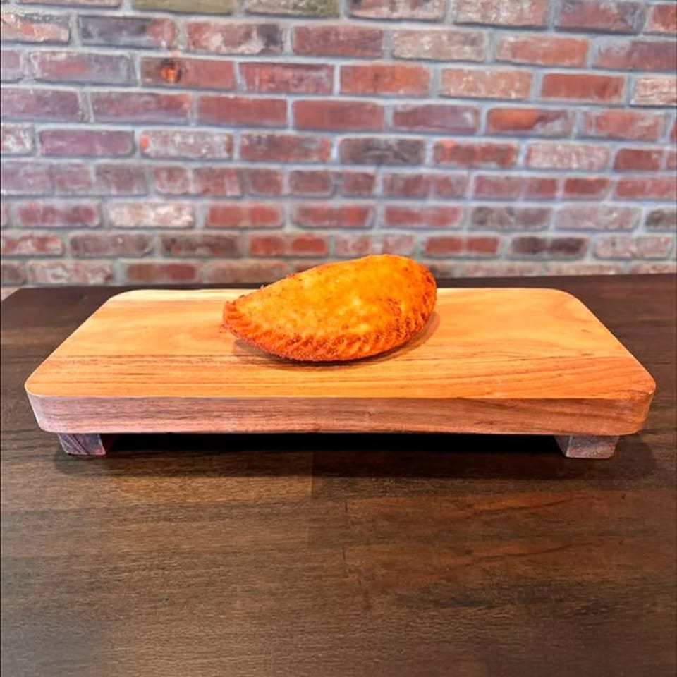 Golden-brown empanada on a wooden serving board, with a brick wall in the background.