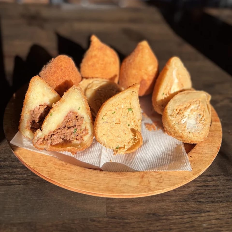 A wooden plate holds several coxinha, some cut open to reveal chicken and beef fillings.