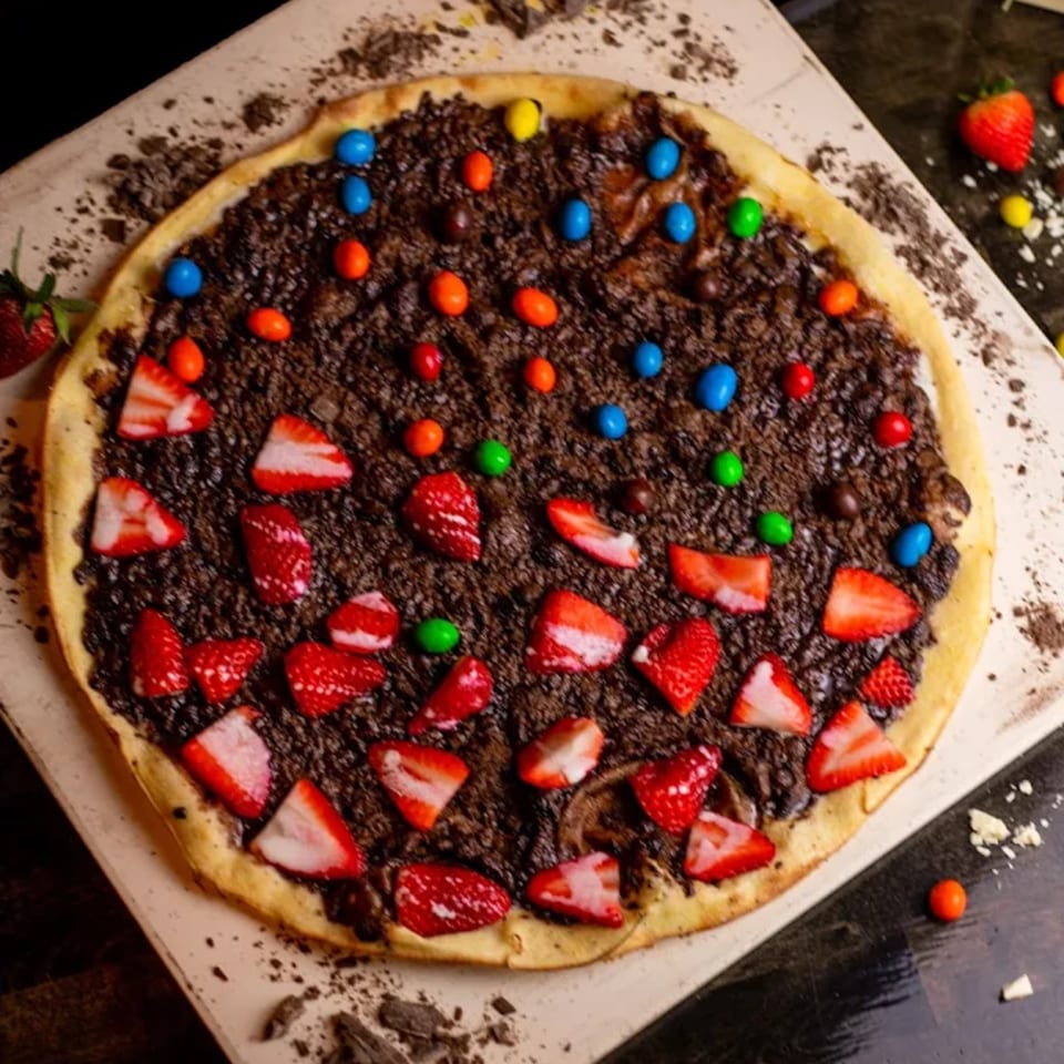 Dessert pizza with chocolate spread, strawberries, and colorful candies on a light-colored surface.